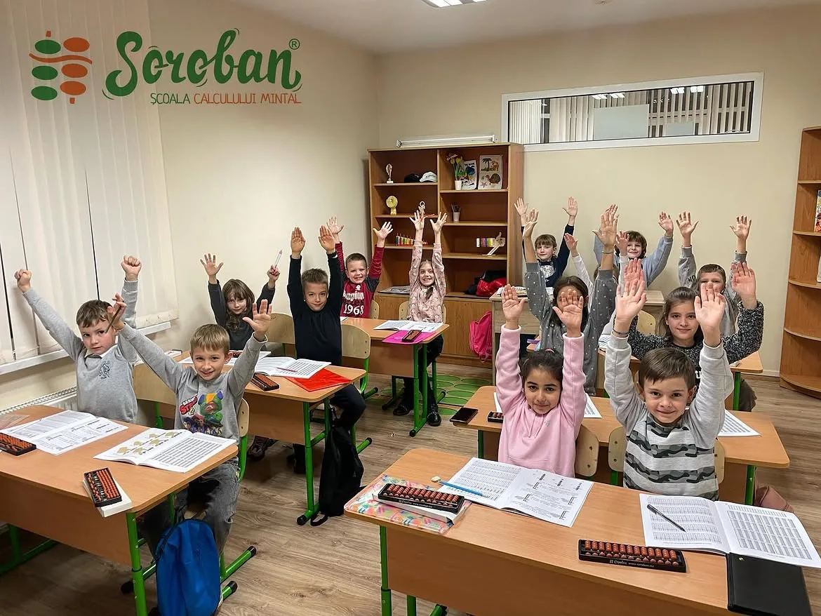 Класс школы SOROBAN: группа детей за партами со счётами и рабочими тетрадями поднимает руки, на стене логотип Soroban